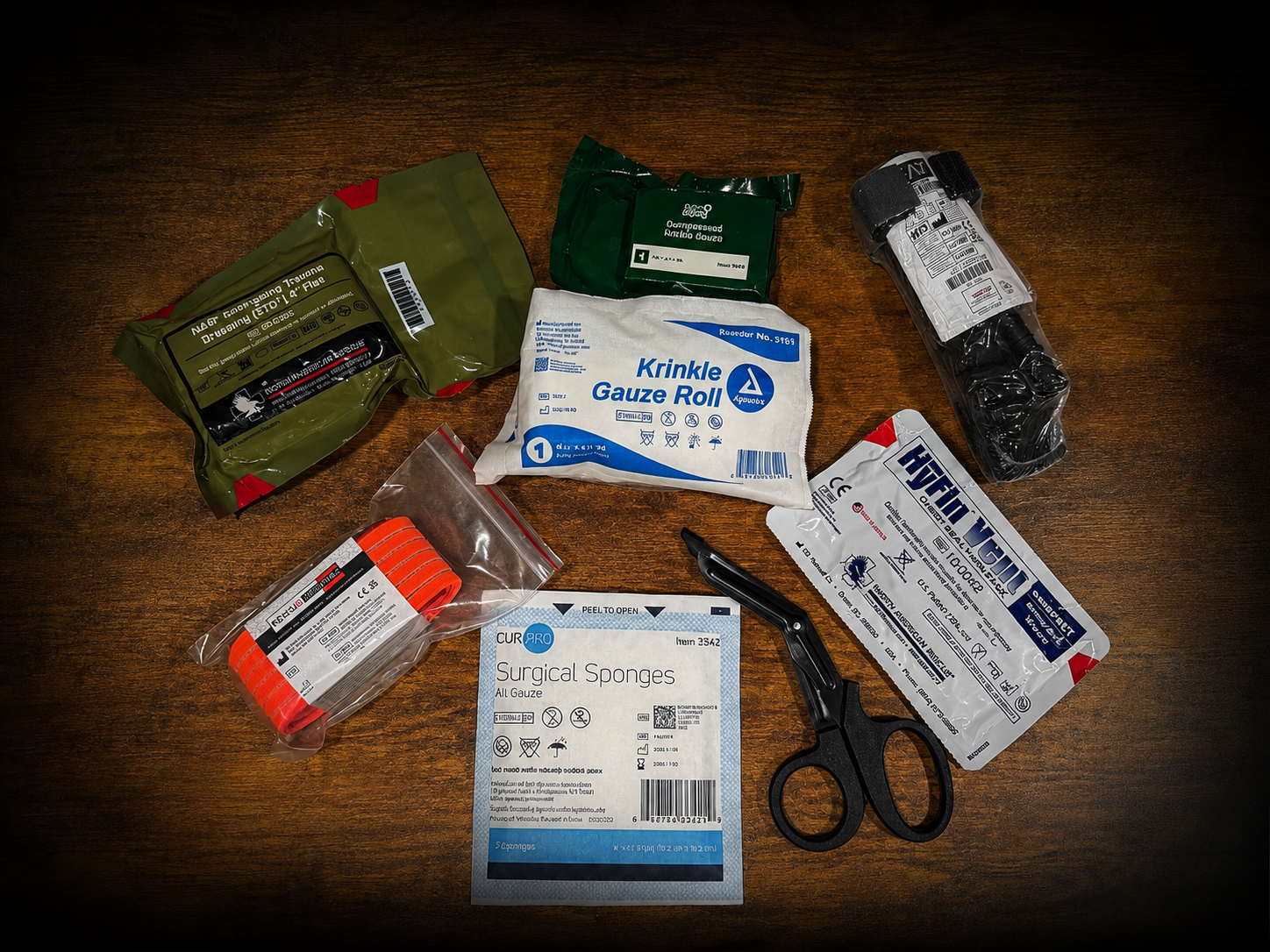 A collection of medical emergency supplies and first aid items arranged on a dark wooden surface, including a black tourniquet, a packaged gauze roll, a tube of hemostatic gauze, a packet of surgical sponges, and other medical kits.