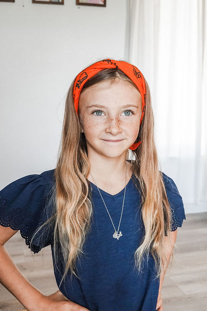 Young girl wearing an orange Multi tool headband and a blue dress indoors.